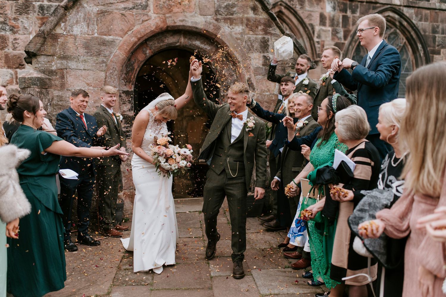 Wedding guests celebrate by blowing bubbles outside a rustic brick building during a joyful outdoor celebration.