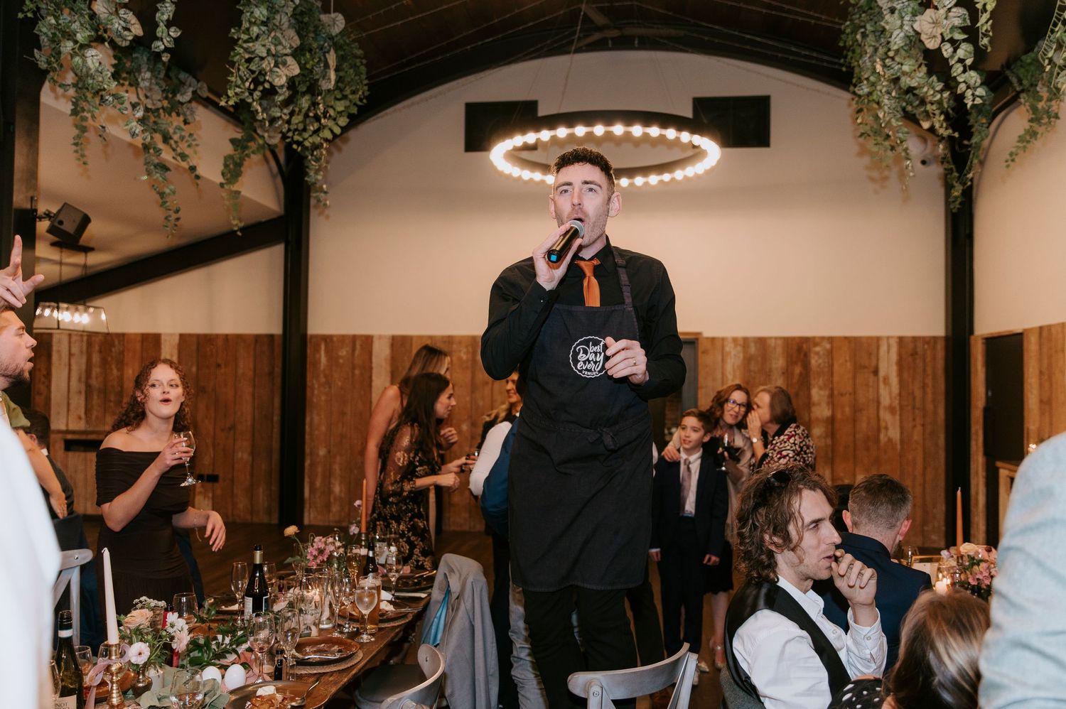Wedding guests gathered in a rustic reception venue with wooden walls and elegant chandelier lighting.
