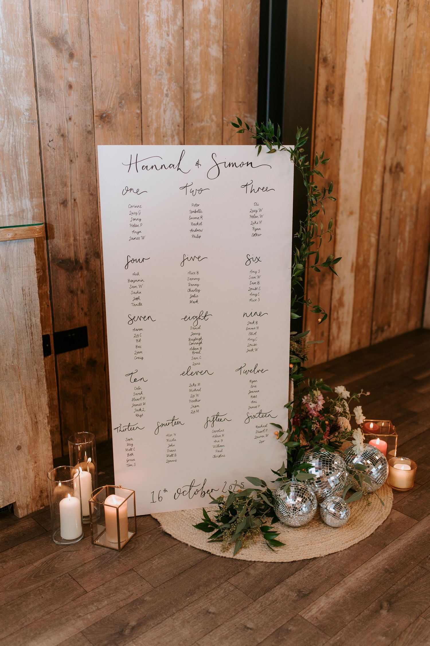 A rustic wedding seating chart display with handwritten text on white boards, decorated with candles and greenery.