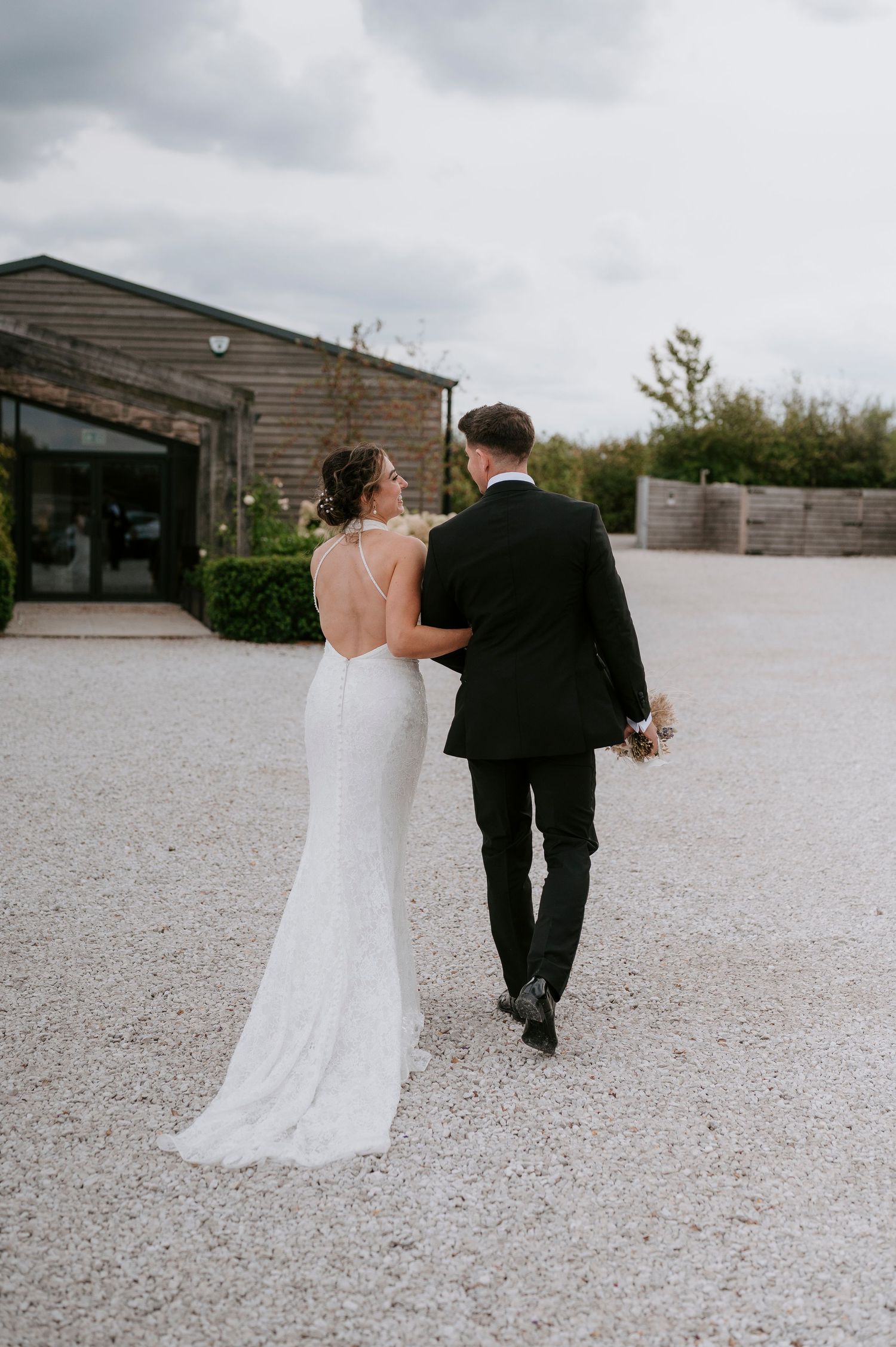 A couple in wedding attire walk away from the camera across a gravel courtyard on their special day.