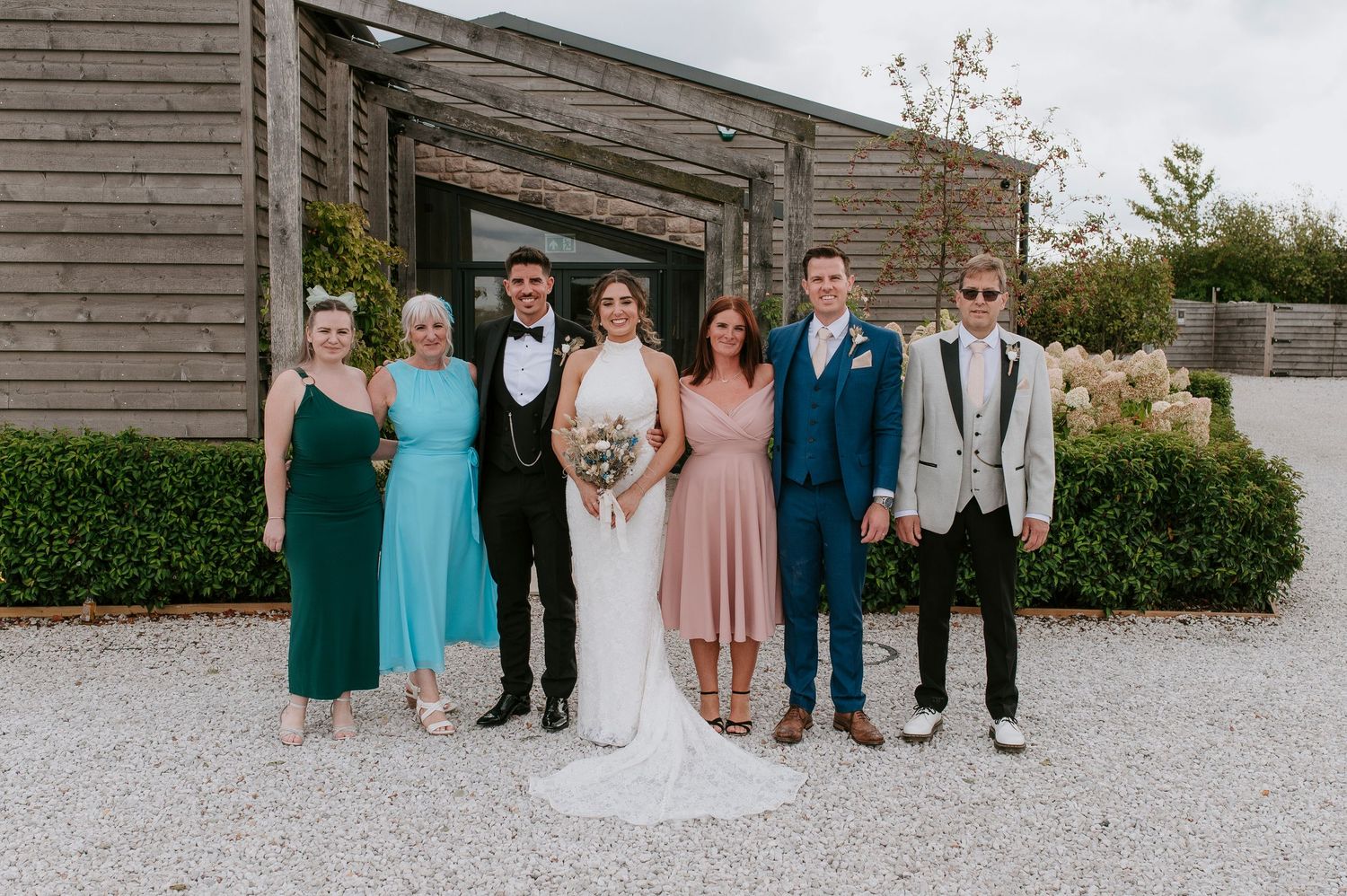 A group of people in formal attire pose together on a gravel path outside a rustic wooden building.