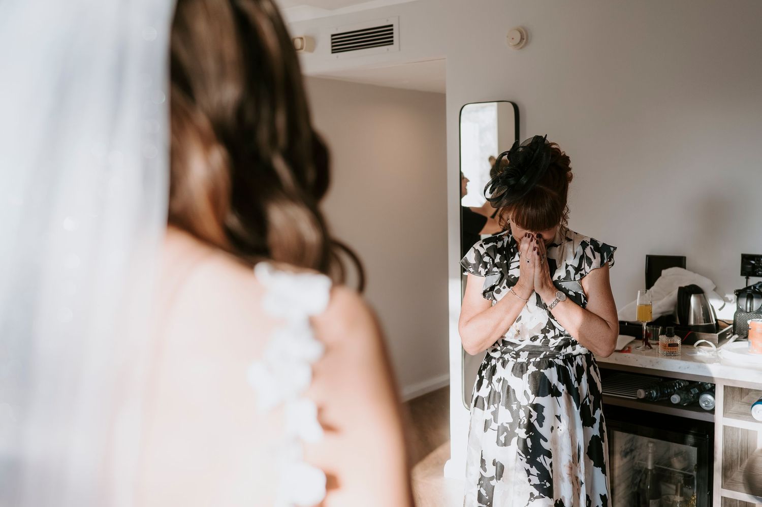 A mother reacts emotionally with hands clasped as she sees her daughter in a wedding dress during bridal preparations.