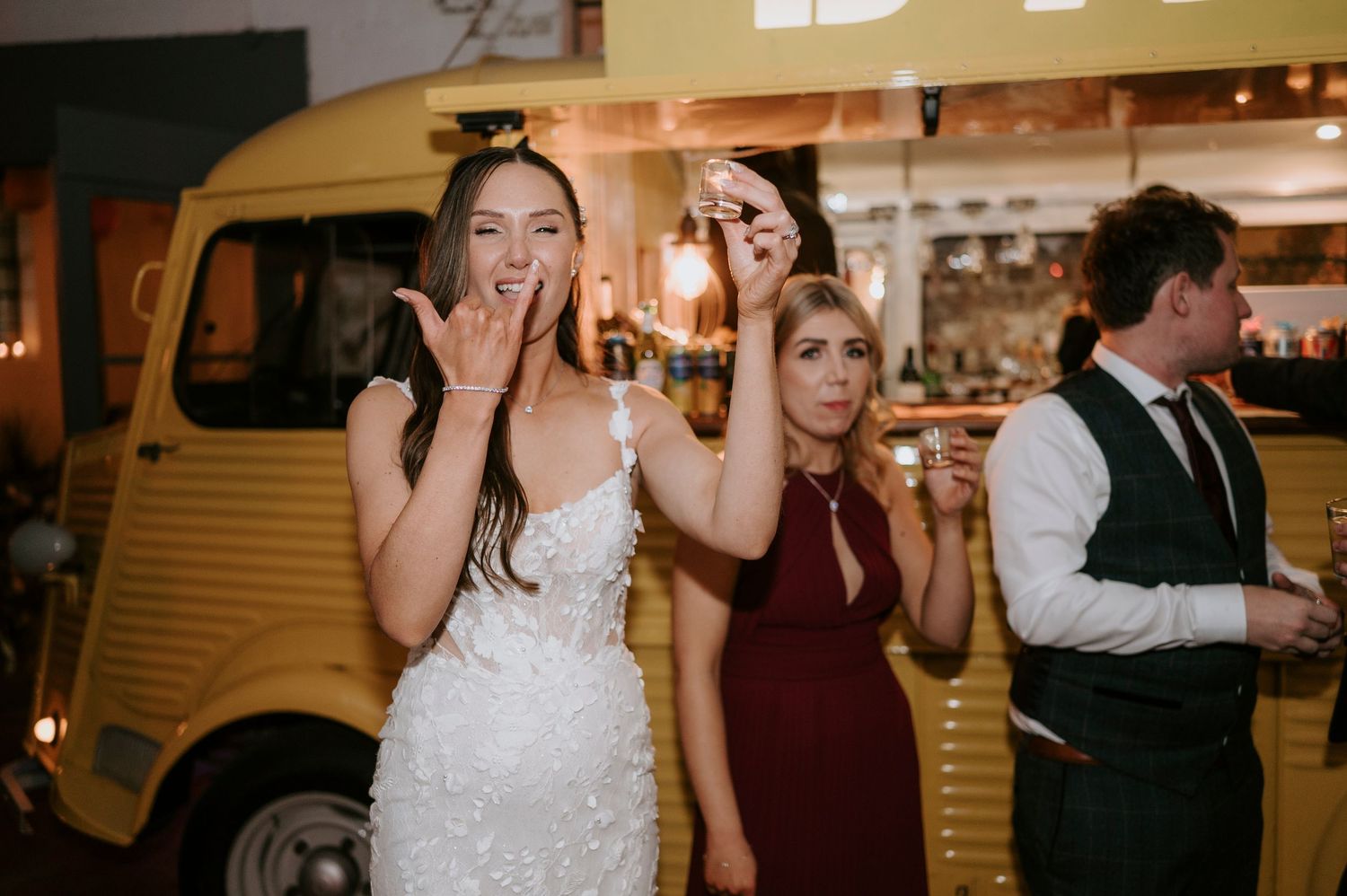Bride in beaded gown poses excitedly with guests near vintage yellow food truck at wedding.