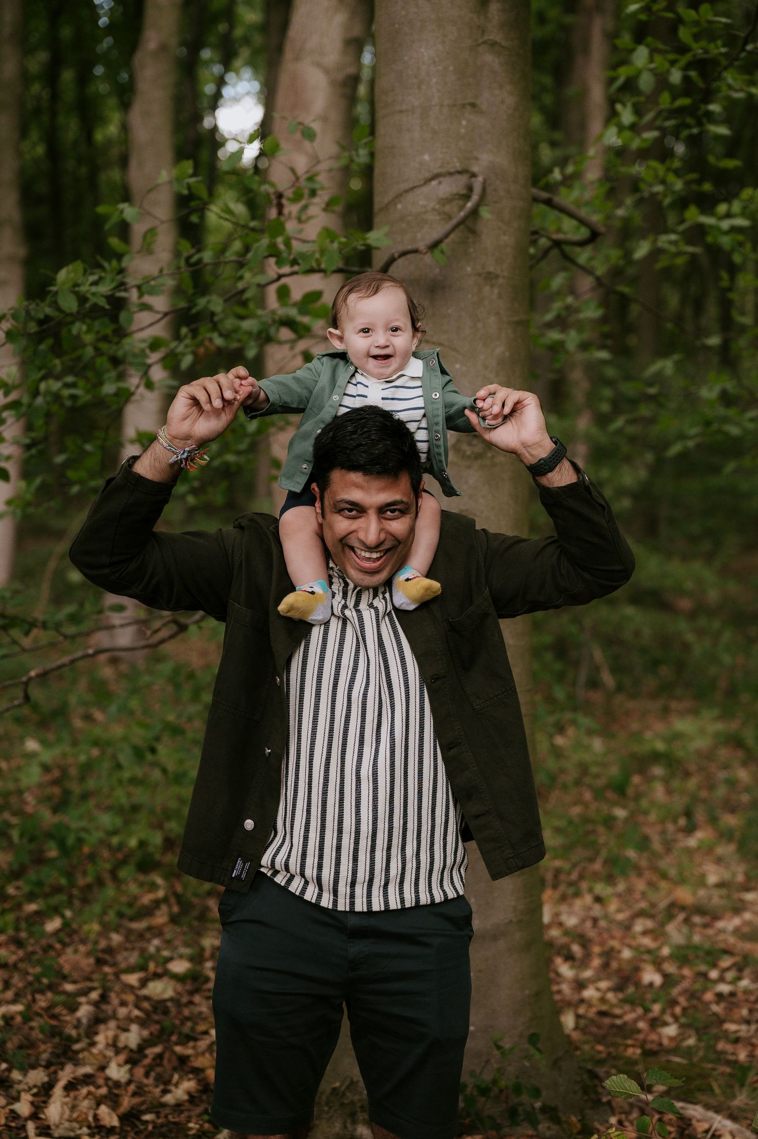 A parent in a black jacket and striped shirt lifts a small child onto their shoulders in a wooded forest setting.