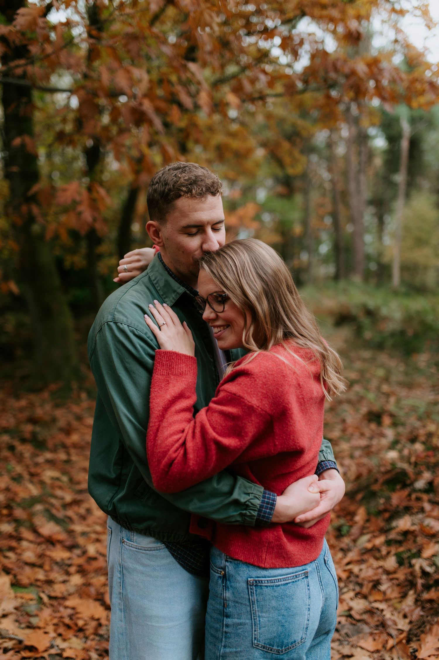 Two people share an intimate moment surrounded by orange fall foliage in a wooded setting.
