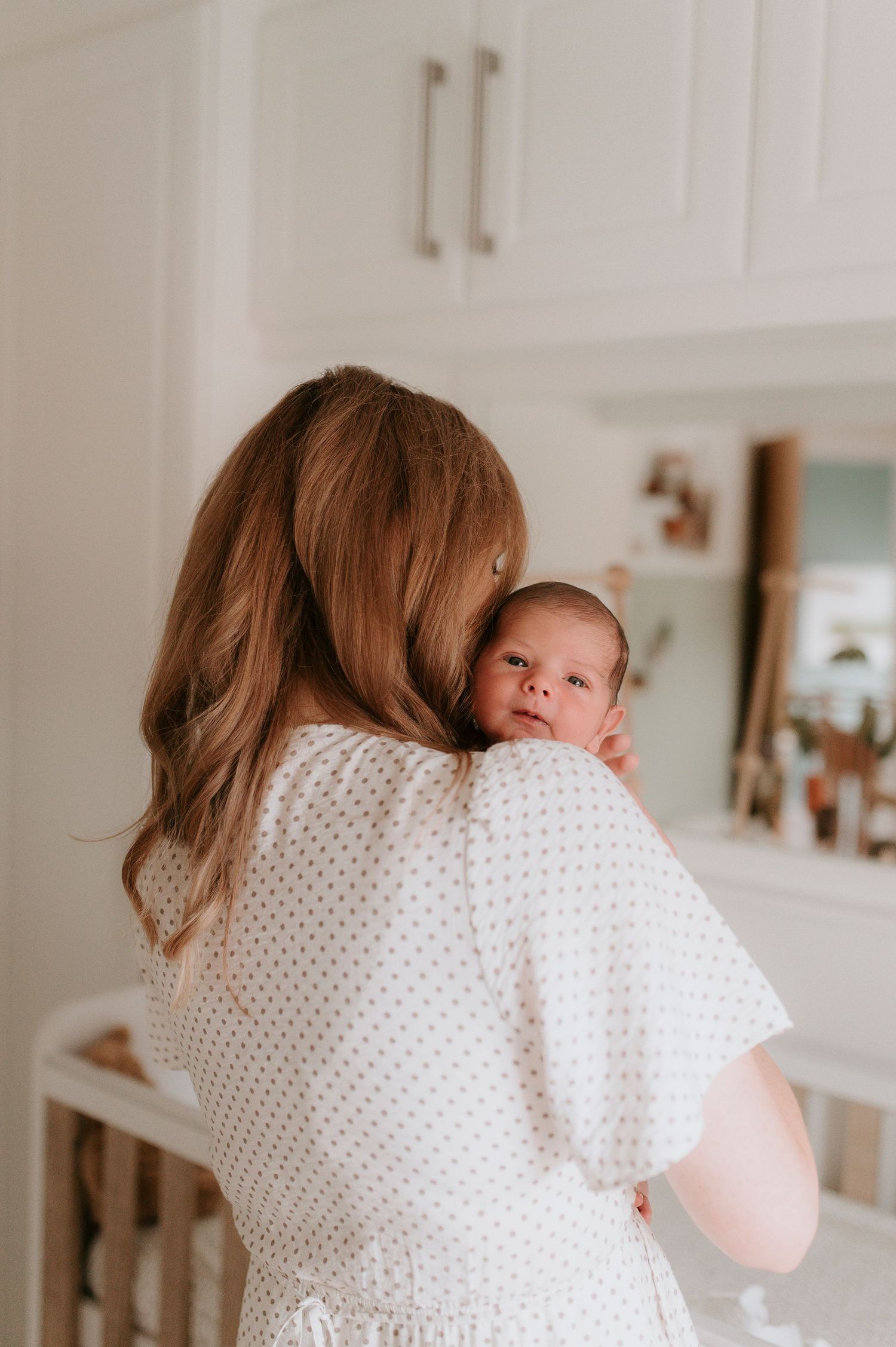 A newborn baby peeks over someone's shoulder wearing a white polka dot dress in a nursery setting.
