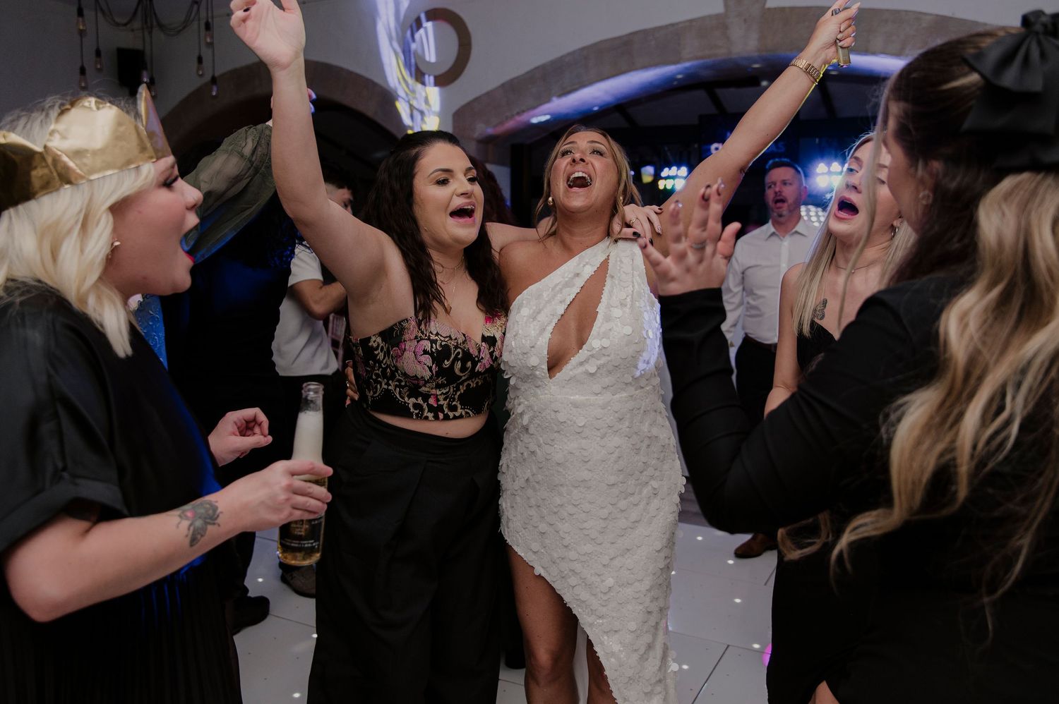 Wedding party guests dancing and celebrating with raised arms on a dimly lit dance floor.
