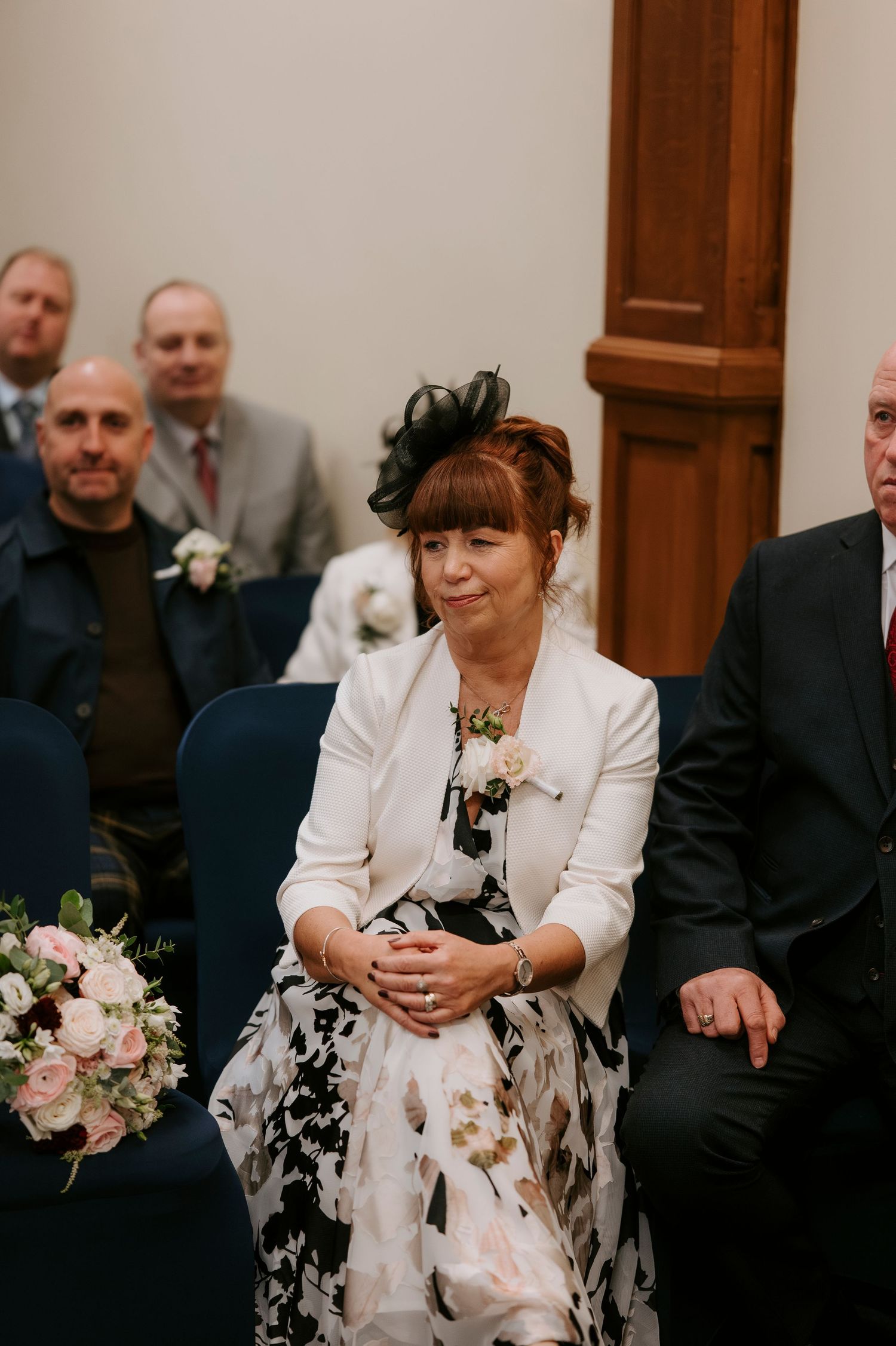 Woman in floral dress and fascinator smiling emotionally at a wedding ceremony.