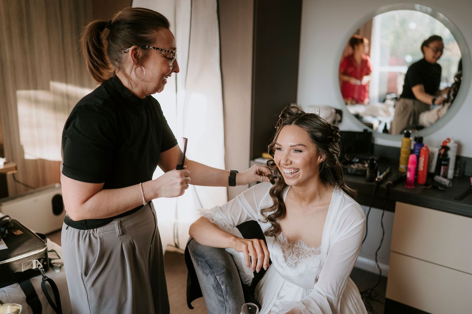 A hairstylist styling a smiling bride's hair in a bright dressing room before her wedding day.