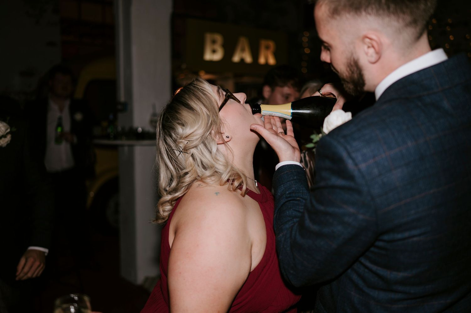Wedding guest in burgundy dress takes a shot at the bar during wedding reception celebration.