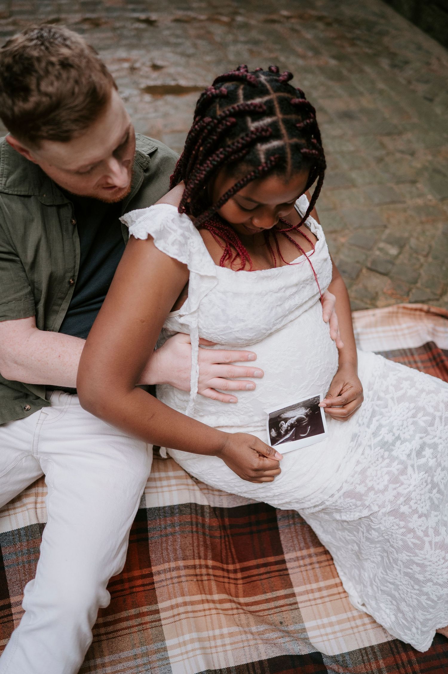 A couple in wedding attire shares a tender moment while signing documents on a plaid blanket outdoors.