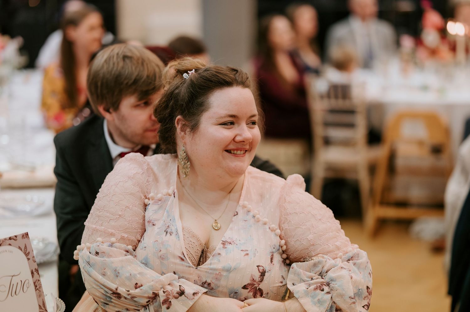 A person in a pink floral dress smiles warmly while seated at a wedding reception.