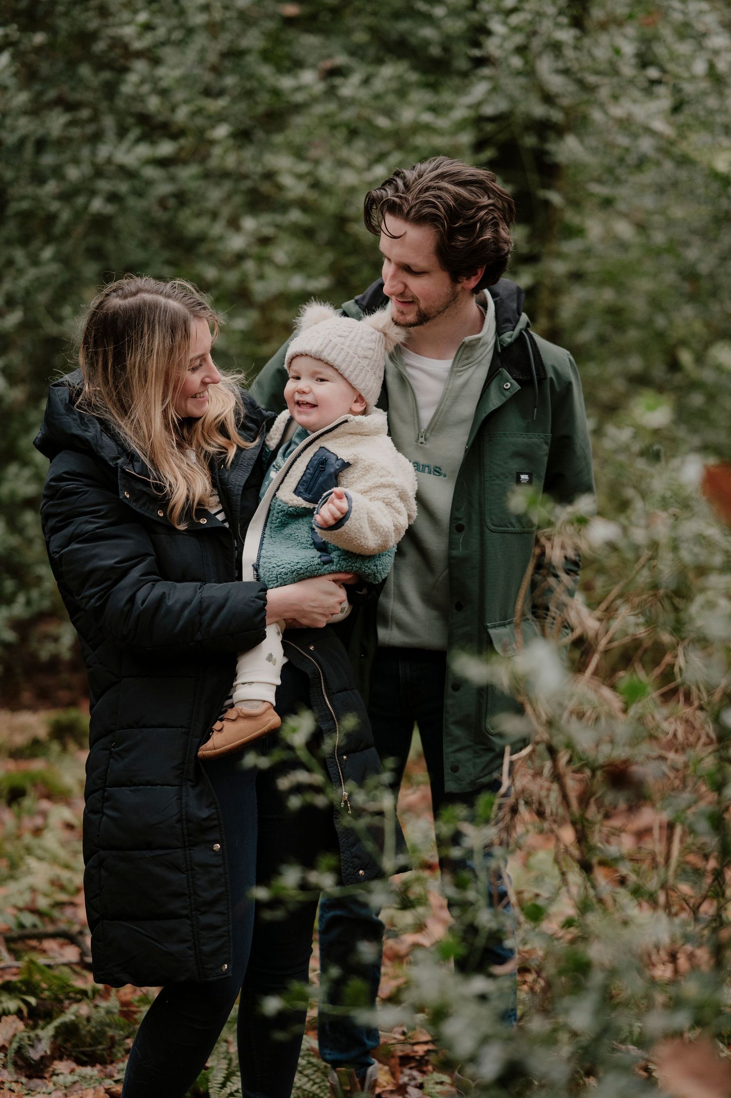 A family enjoys a tender moment together outdoors in autumn, surrounded by greenery and wearing warm winter coats.