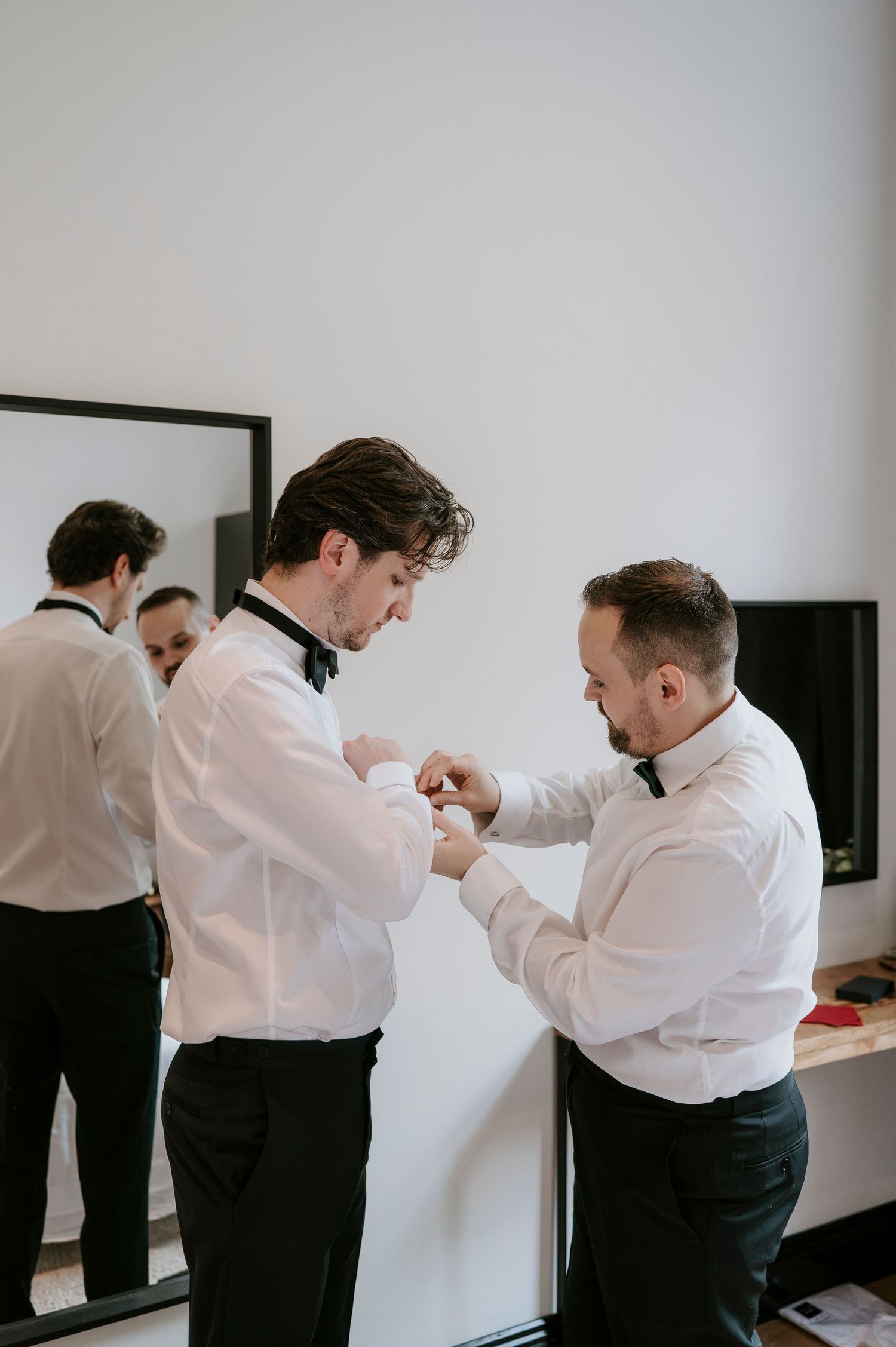 Groomsmen in white shirts and black pants helping each other get ready before the wedding ceremony.