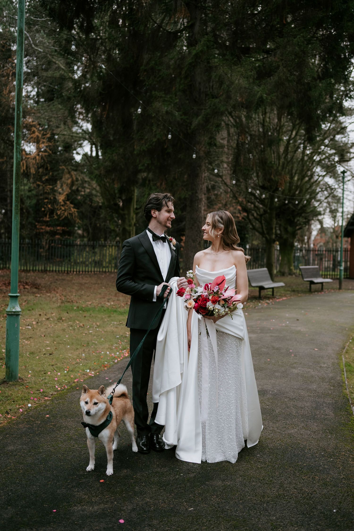 A couple in formal attire walks with their Shiba Inu dog along a tree-lined path in a park setting.