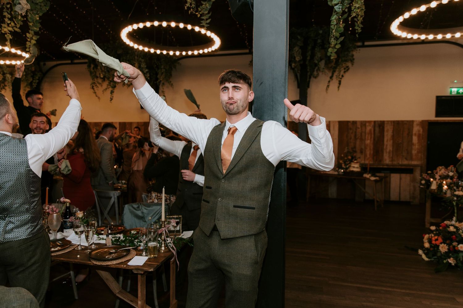 Wedding reception servers in vests carry plates of food through an elegantly decorated venue with string lights overhead.