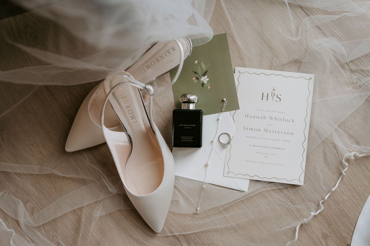 Elegant wedding details including white invitations, delicate lace veil, and blush pink heels arranged on light surface.