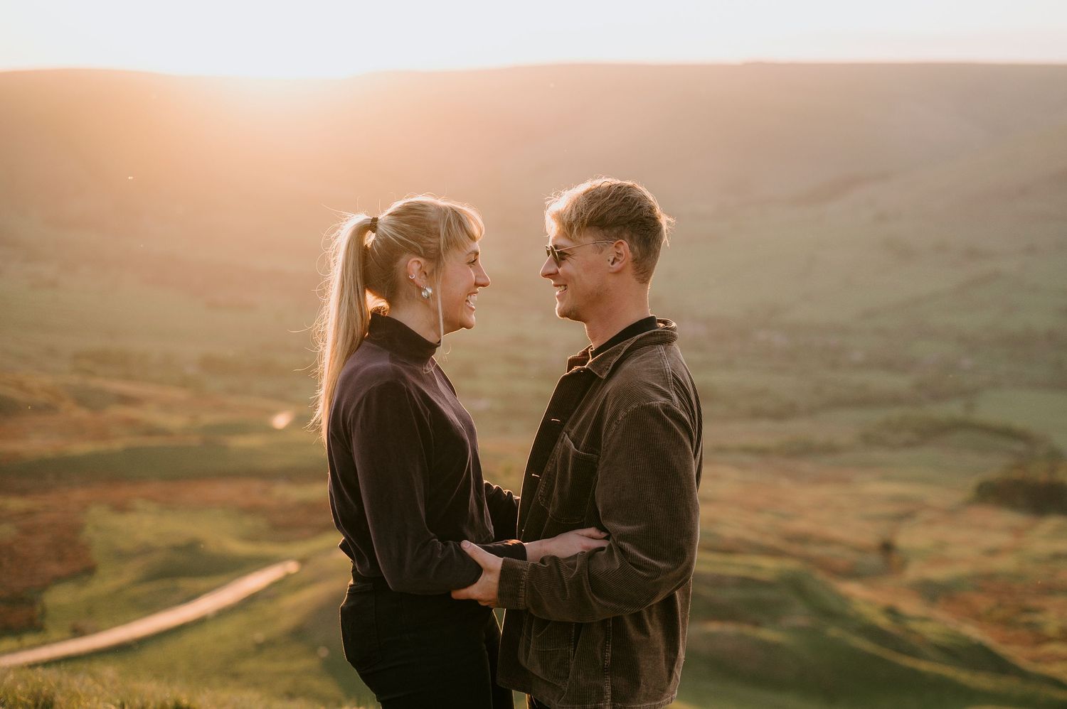A couple stands close together on a scenic hilltop overlooking a misty rural landscape at sunset.