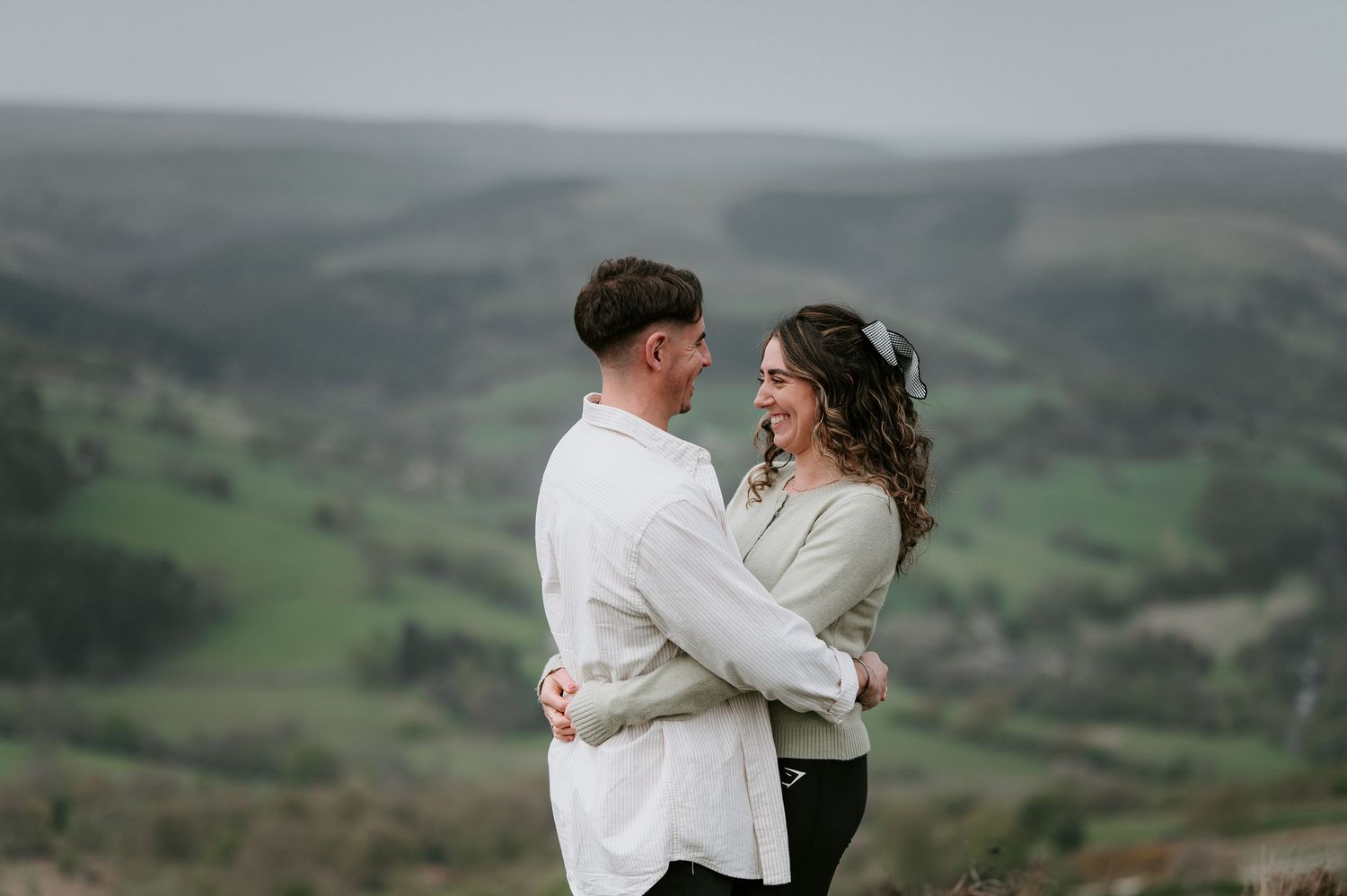 A couple in white embraces while overlooking a scenic green mountain landscape on an overcast day.