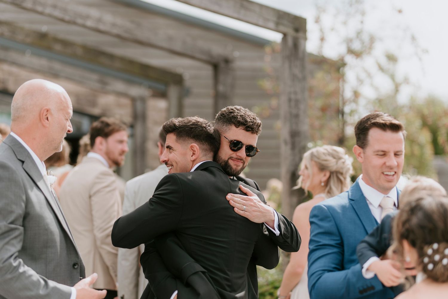Wedding guests share emotional embraces and greetings during an outdoor celebration at a stone venue.