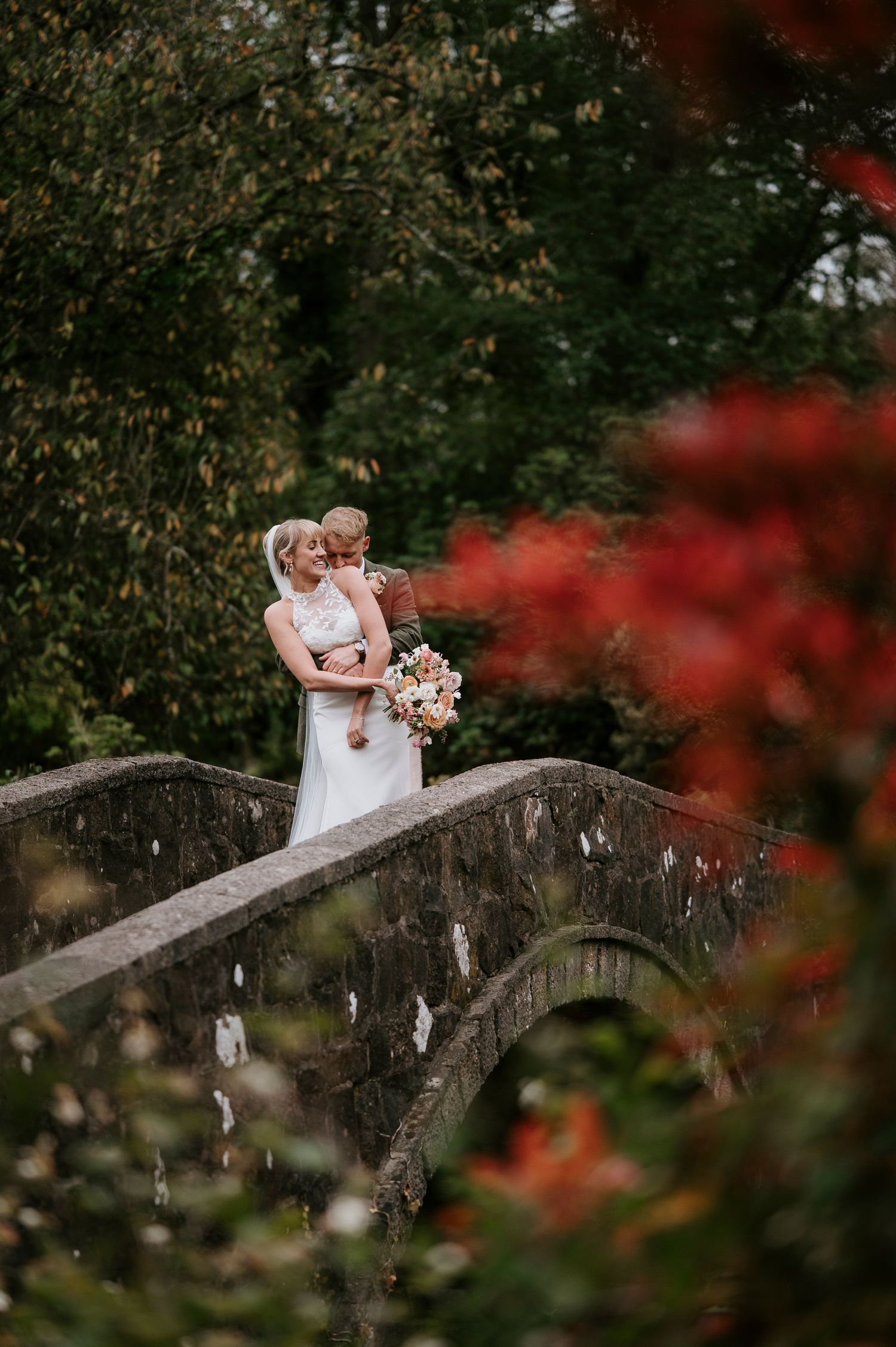 A couple shares an intimate moment on a rustic stone bridge surrounded by autumn foliage and red flowers in the foreground.