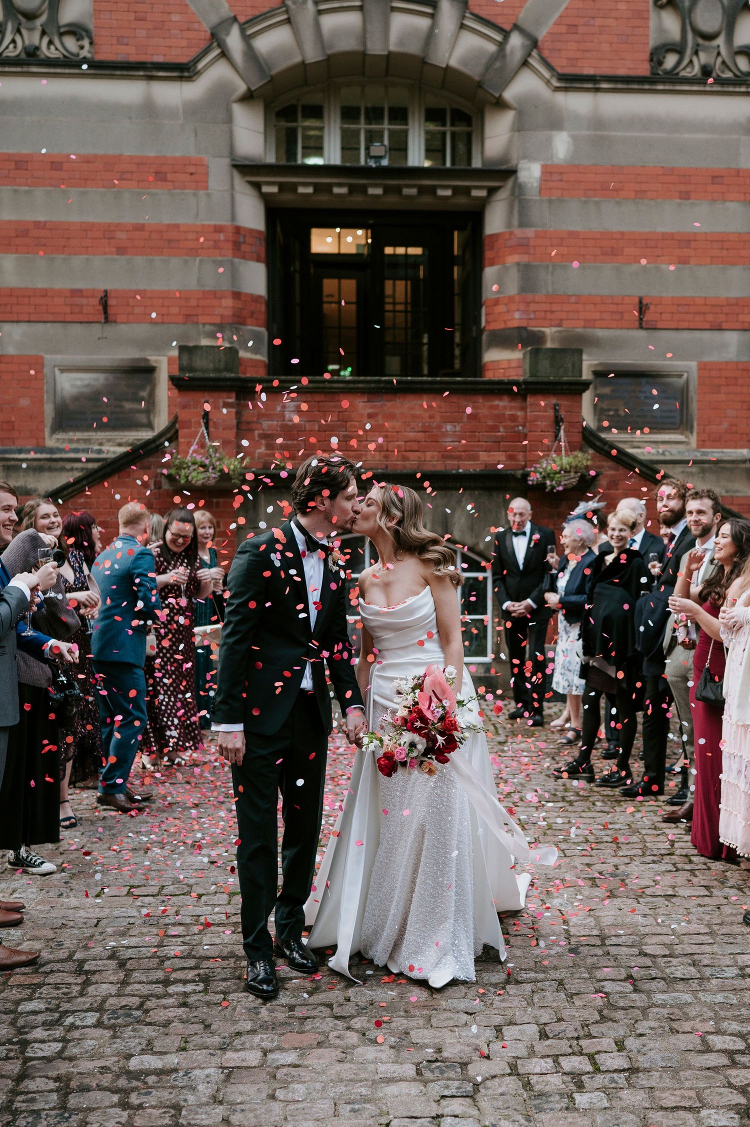 Wedding party and guests showered with confetti outside historic brick building with cobblestone path.
