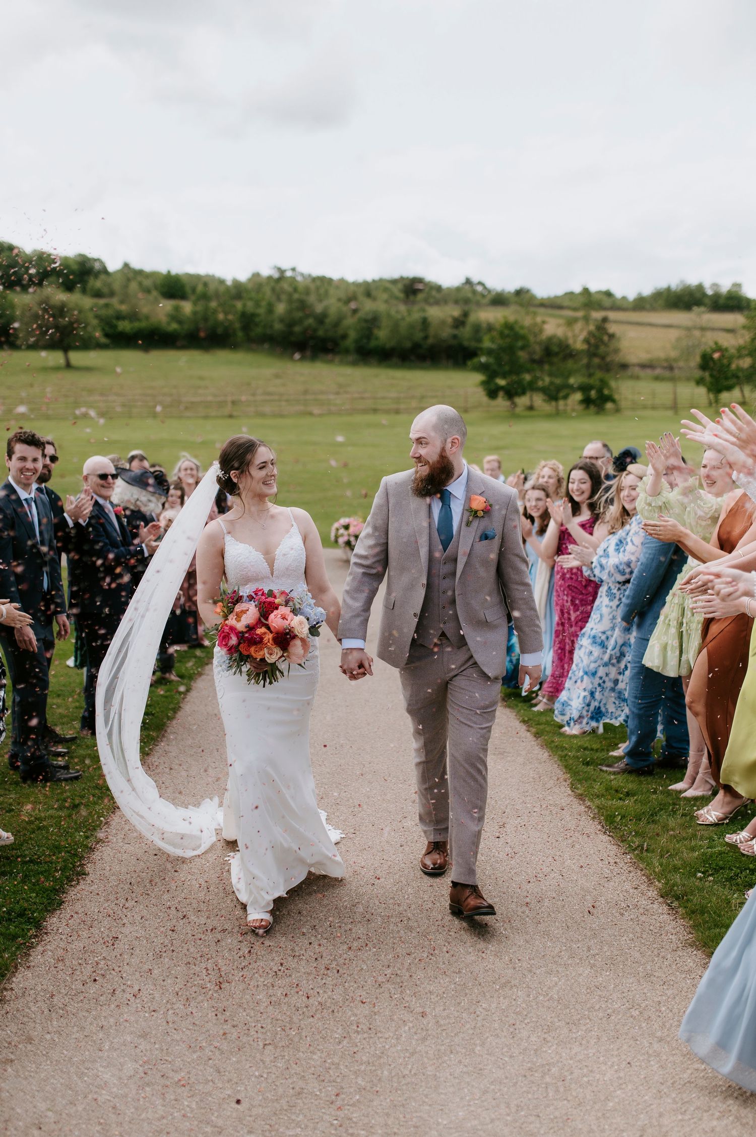 Wedding guests line a path as newlyweds walk between them during an outdoor countryside celebration.