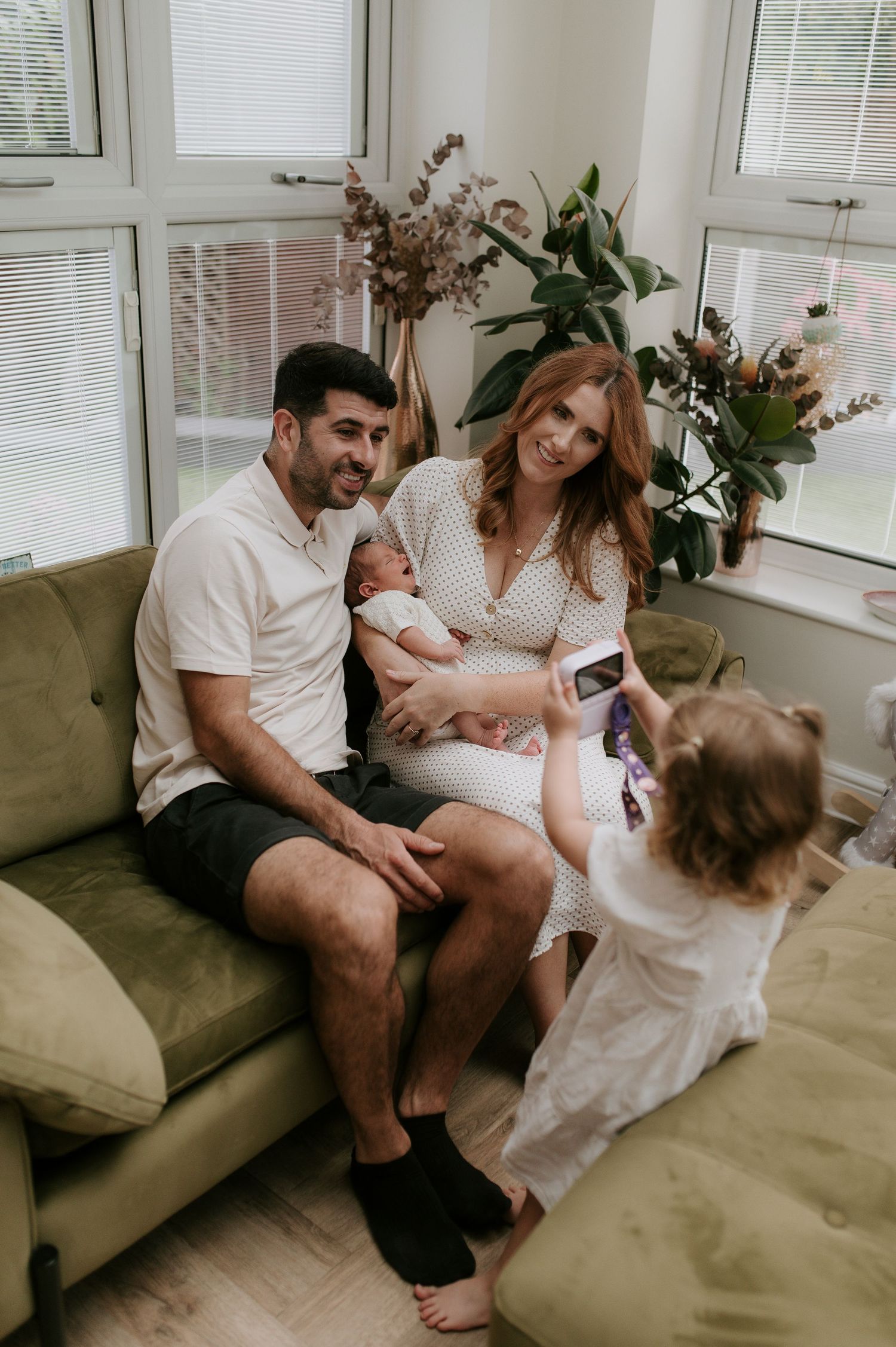 A family shares joyful moments together on a cozy gray couch in a bright, window-lit living room.