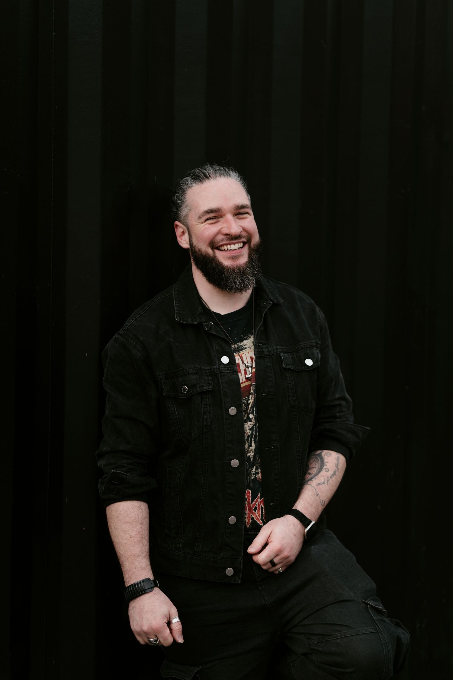 A person in a black denim jacket laughs heartily while seated against a dark background.