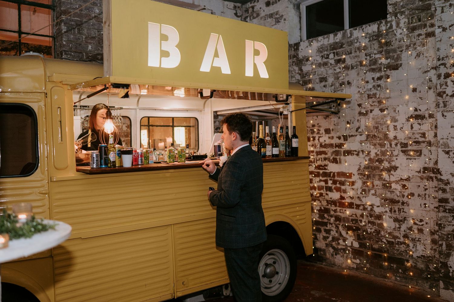 A man orders drinks at a vintage yellow mobile bar truck with illuminated BAR sign at an indoor wedding reception.