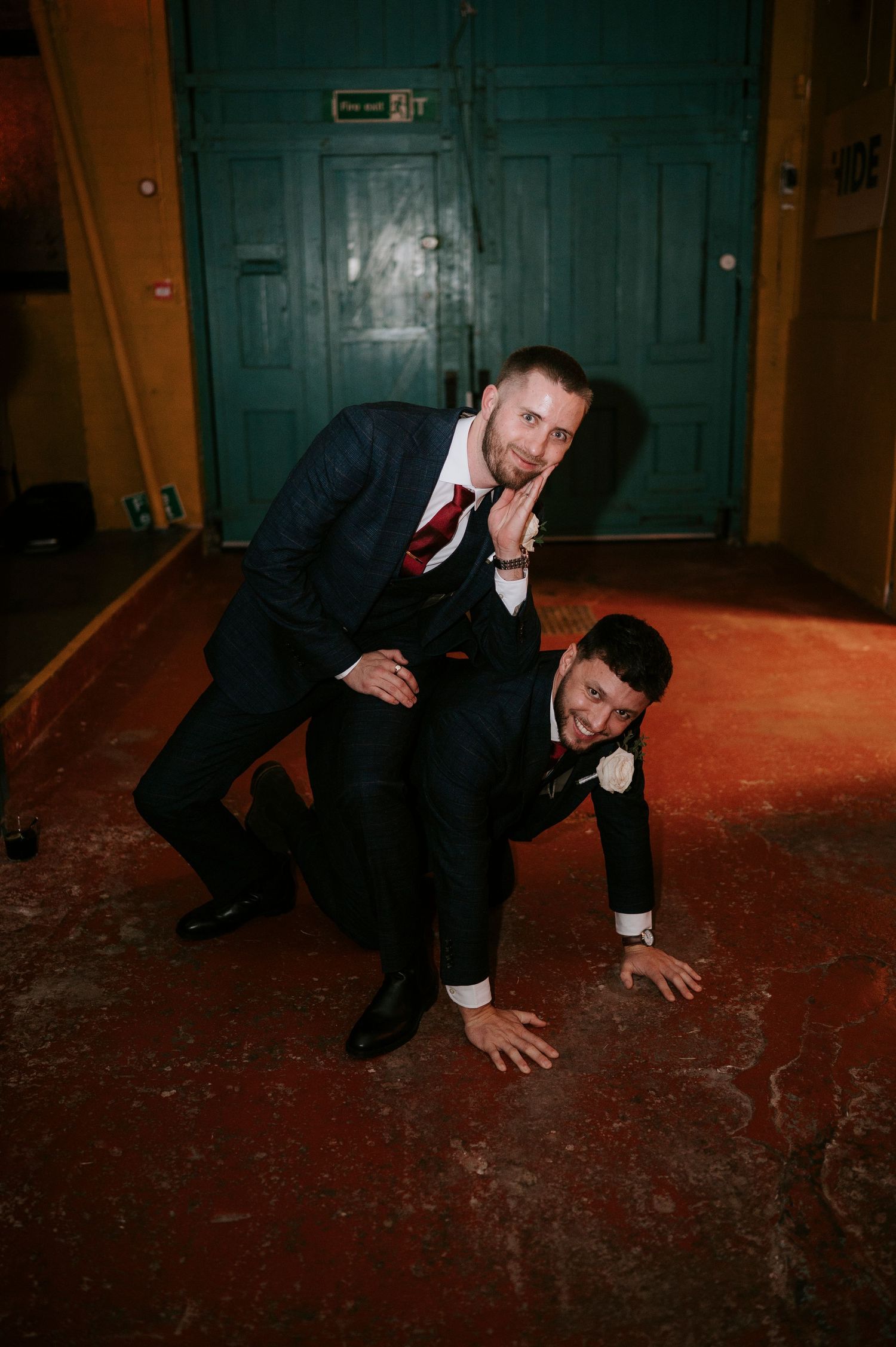 Two groomsmen in navy suits playfully wrestle on the floor at a wedding reception venue.