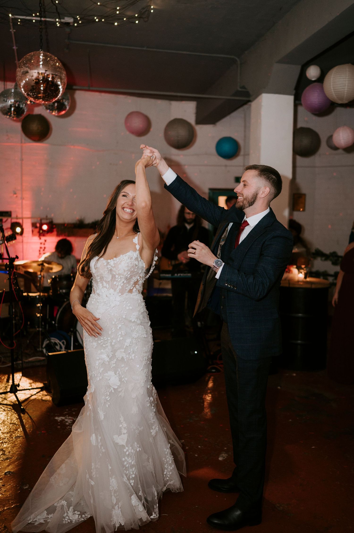 Groom twirls bride in lace wedding gown on dance floor with colorful paper lanterns overhead.
