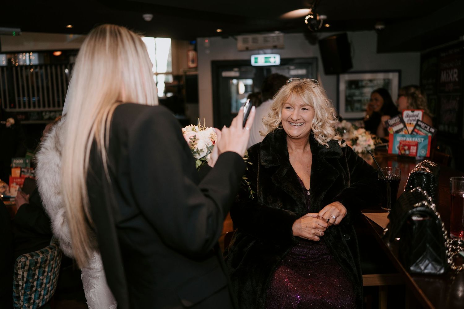 Two women socializing in a bar, one wearing a black blazer with wrist corsage, the other in a velvet outfit.