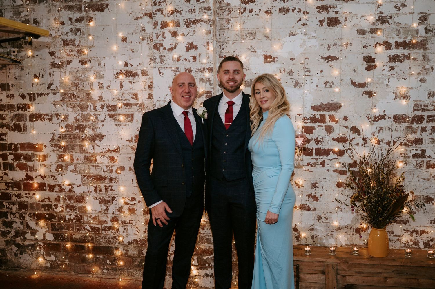 Man and woman in formal attire pose with groom in front of fairy light-adorned exposed brick wall at wedding.