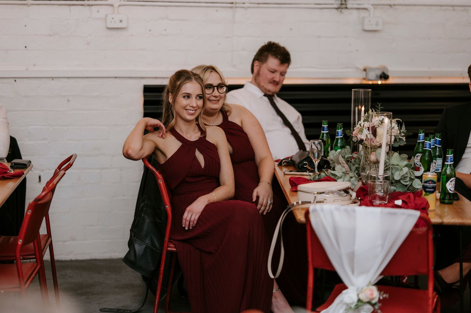 Two bridesmaids in burgundy dresses pose with male guest at wedding reception table with candles.