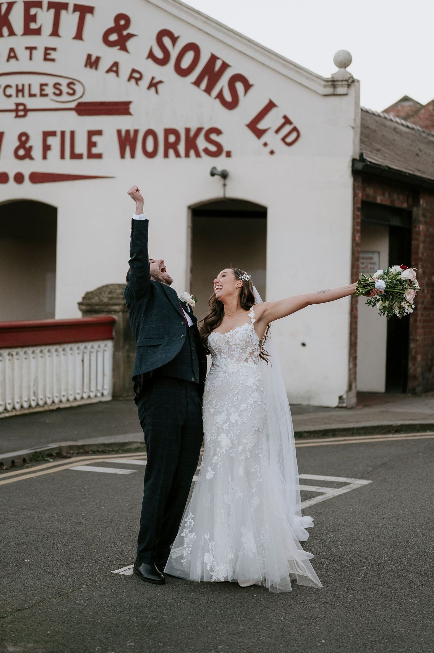 Ecstatic bride and groom pose outside historic Beckett & Sons building, both raising arms in celebration.