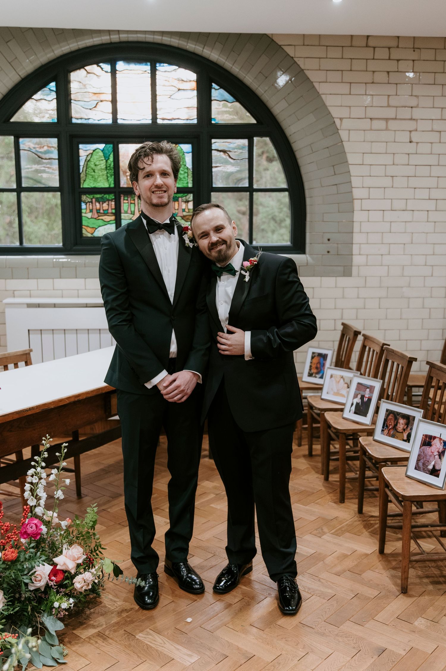 Two people in formal black suits standing in a room with arched windows and wooden chairs.