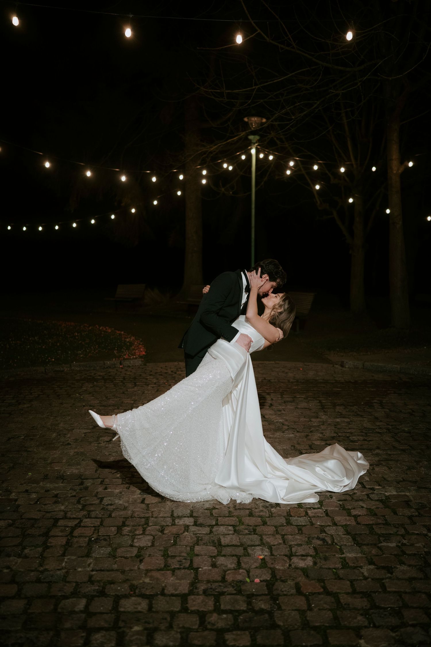 Romantic dance scene under string lights with flowing white wedding dress on cobblestone ground.