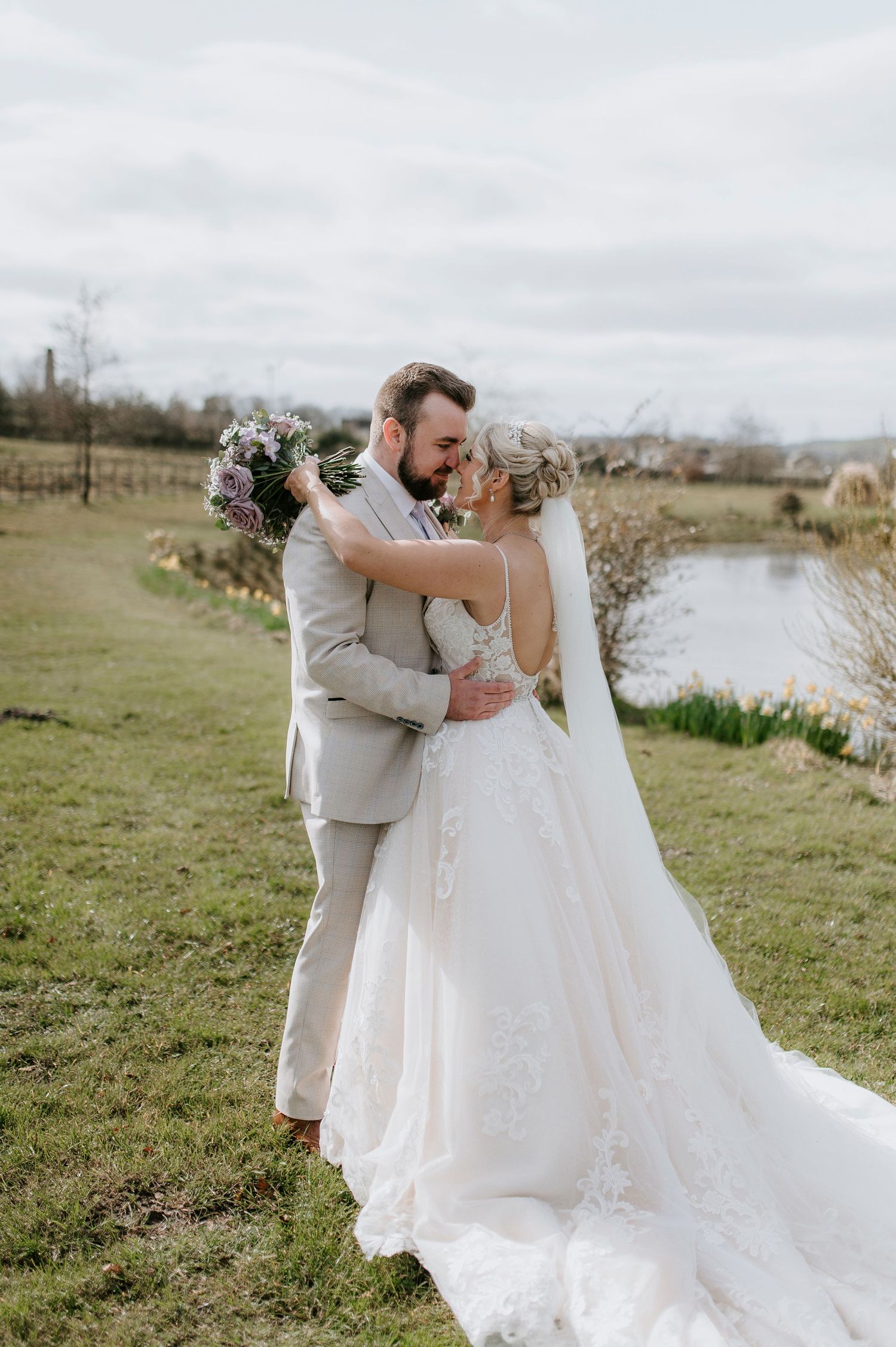 A romantic wedding couple embraces on a grassy hill overlooking a serene pond under cloudy skies.