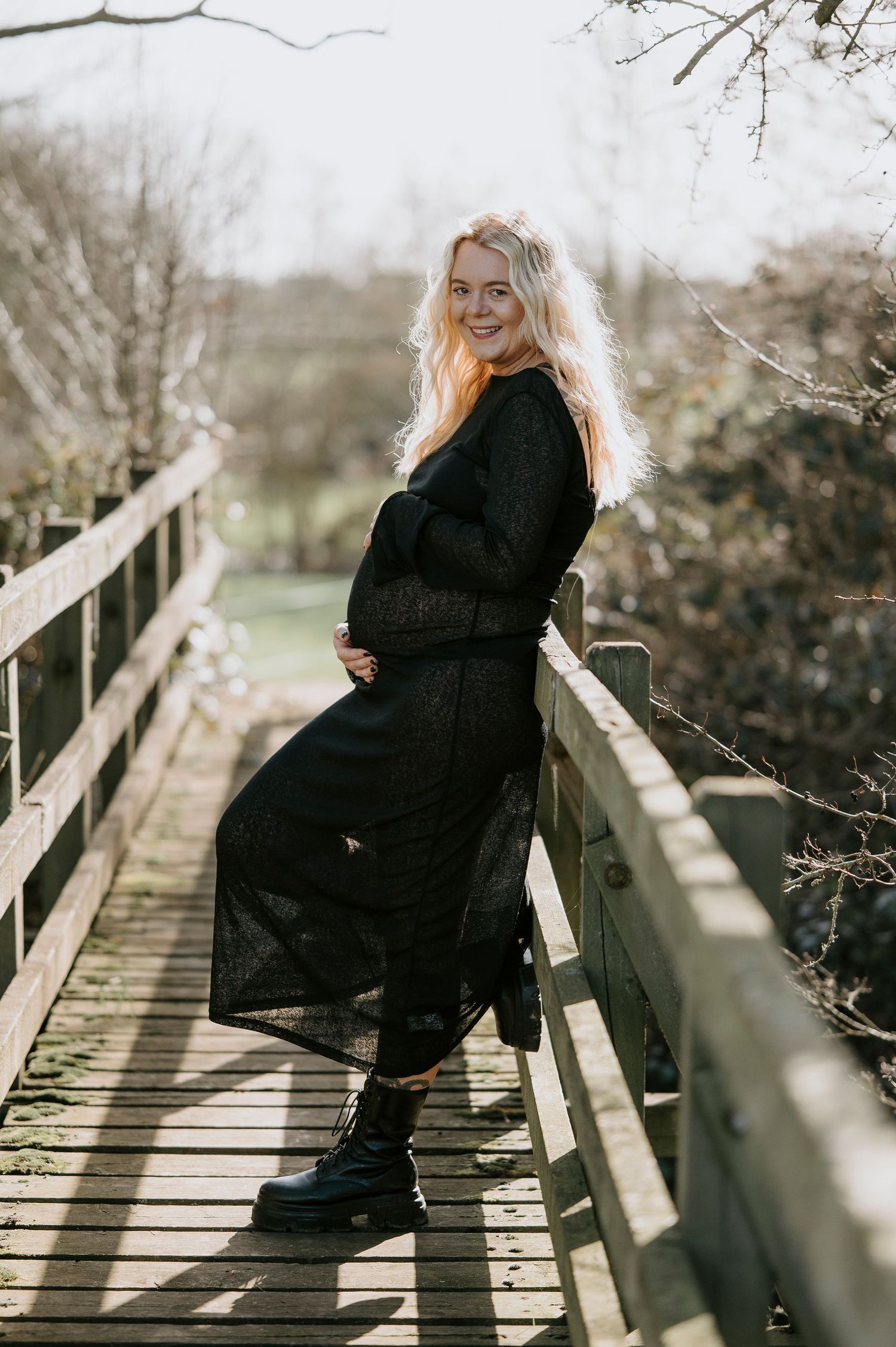 Person in dark clothing poses on rustic wooden bridge surrounded by bare winter branches.
