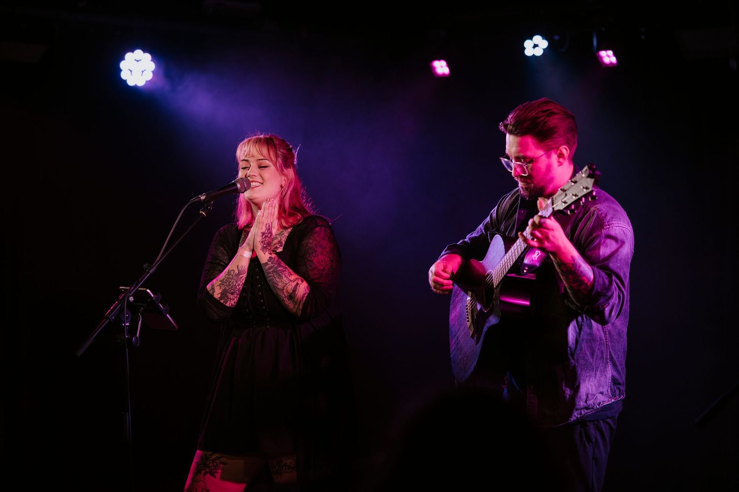 Musicians perform on a dark stage with dramatic purple and blue lighting effects during a live concert performance.