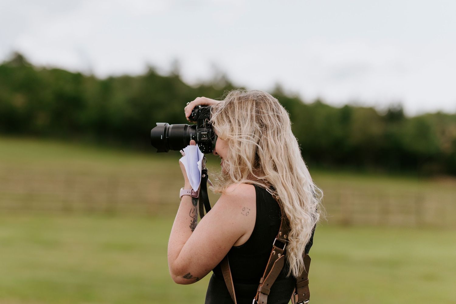 Photographer with long blonde hair takes pictures outdoors in a grassy field during golden hour.