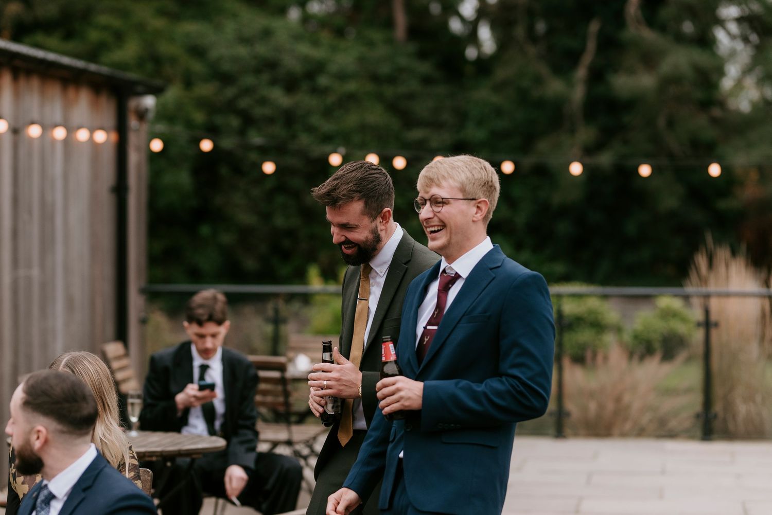 Groomsmen in navy suits clink beer bottles together in celebration at an outdoor wedding reception.