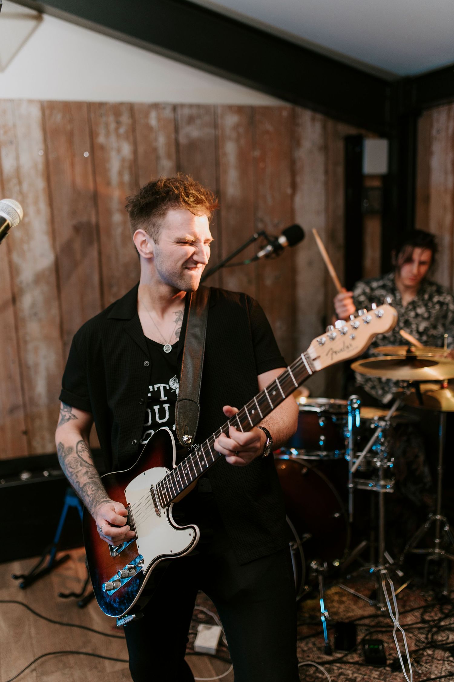 A musician plays an electric guitar during a live performance with band equipment visible in the background.