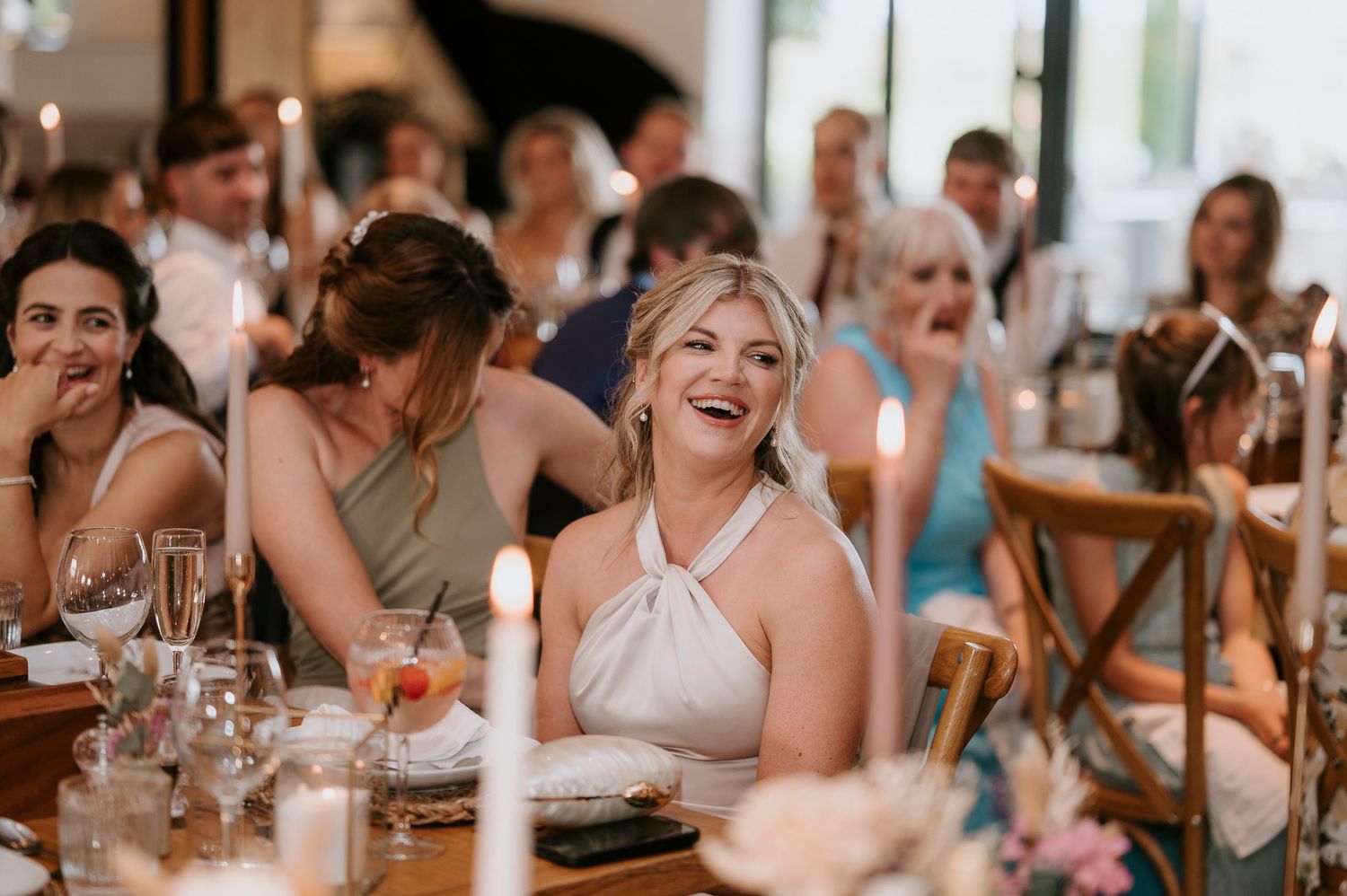 Wedding guests enjoying dinner and conversation at long tables in a bright, airy reception venue.