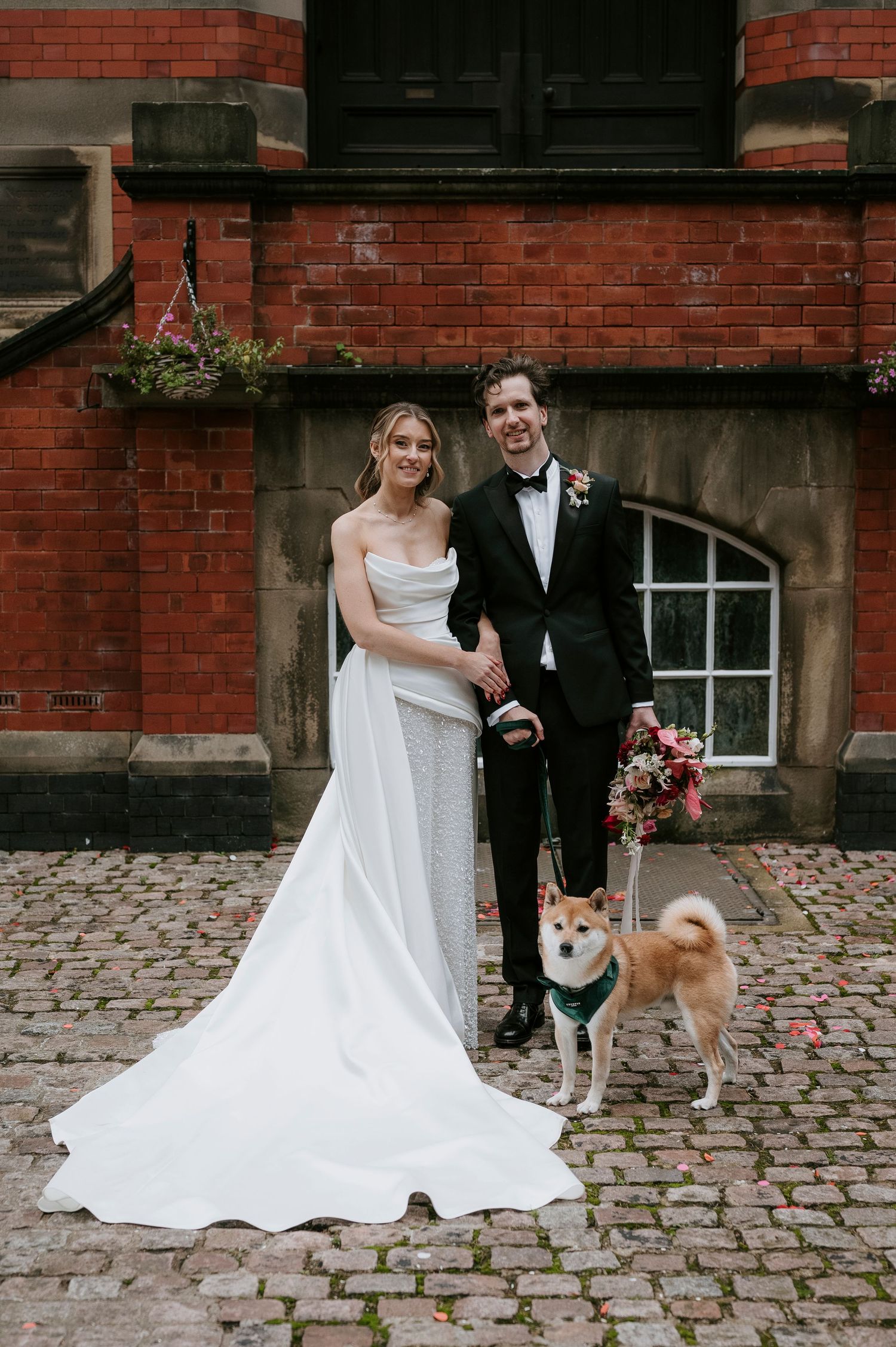 A couple in wedding attire poses with their corgi dog on cobblestone ground against a brick building exterior.