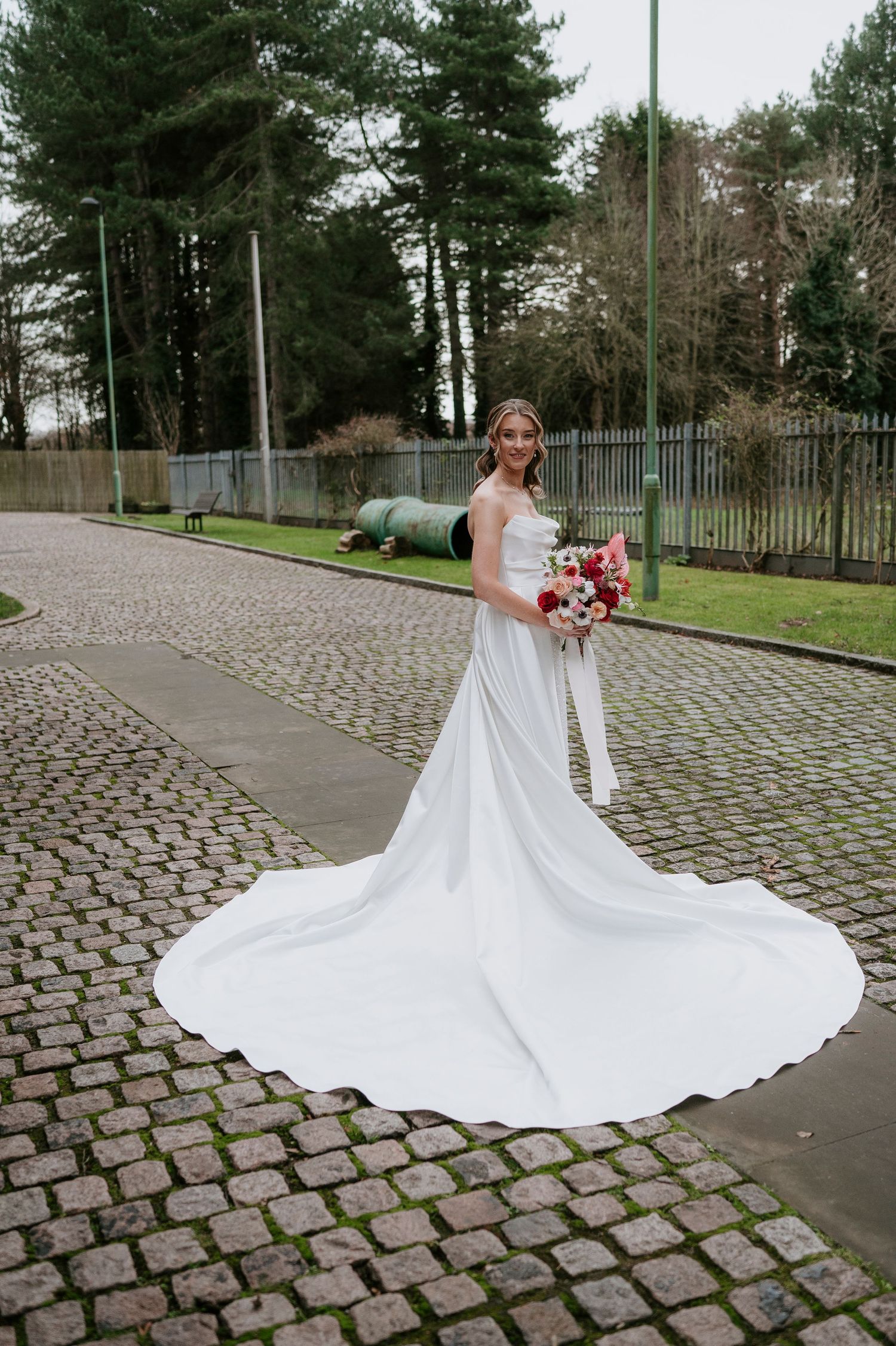White wedding dress with flowing train displayed on cobblestone pavement surrounded by pine trees.