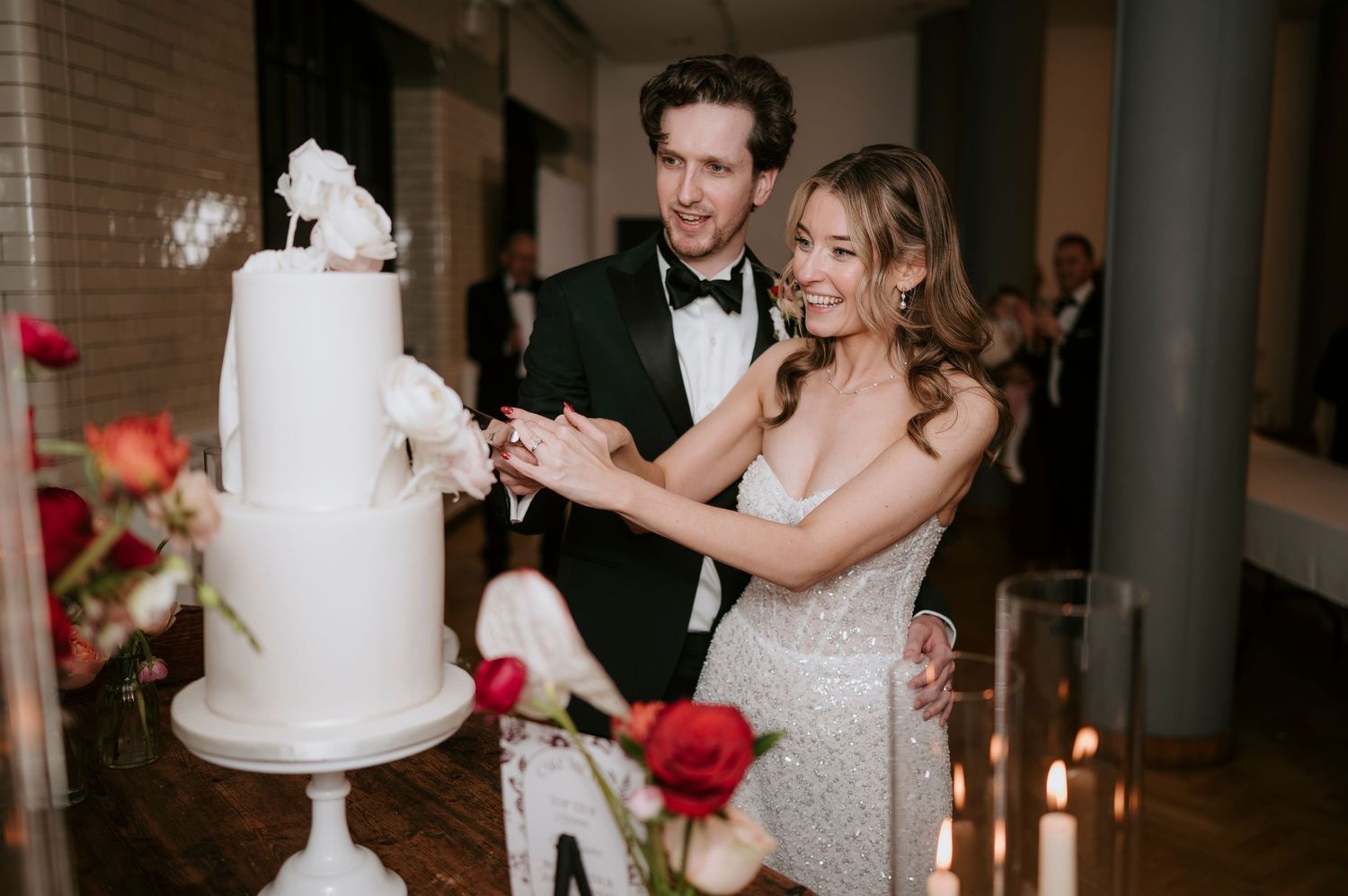 A bride and groom cut into their elegant white wedding cake together at a candlelit reception.