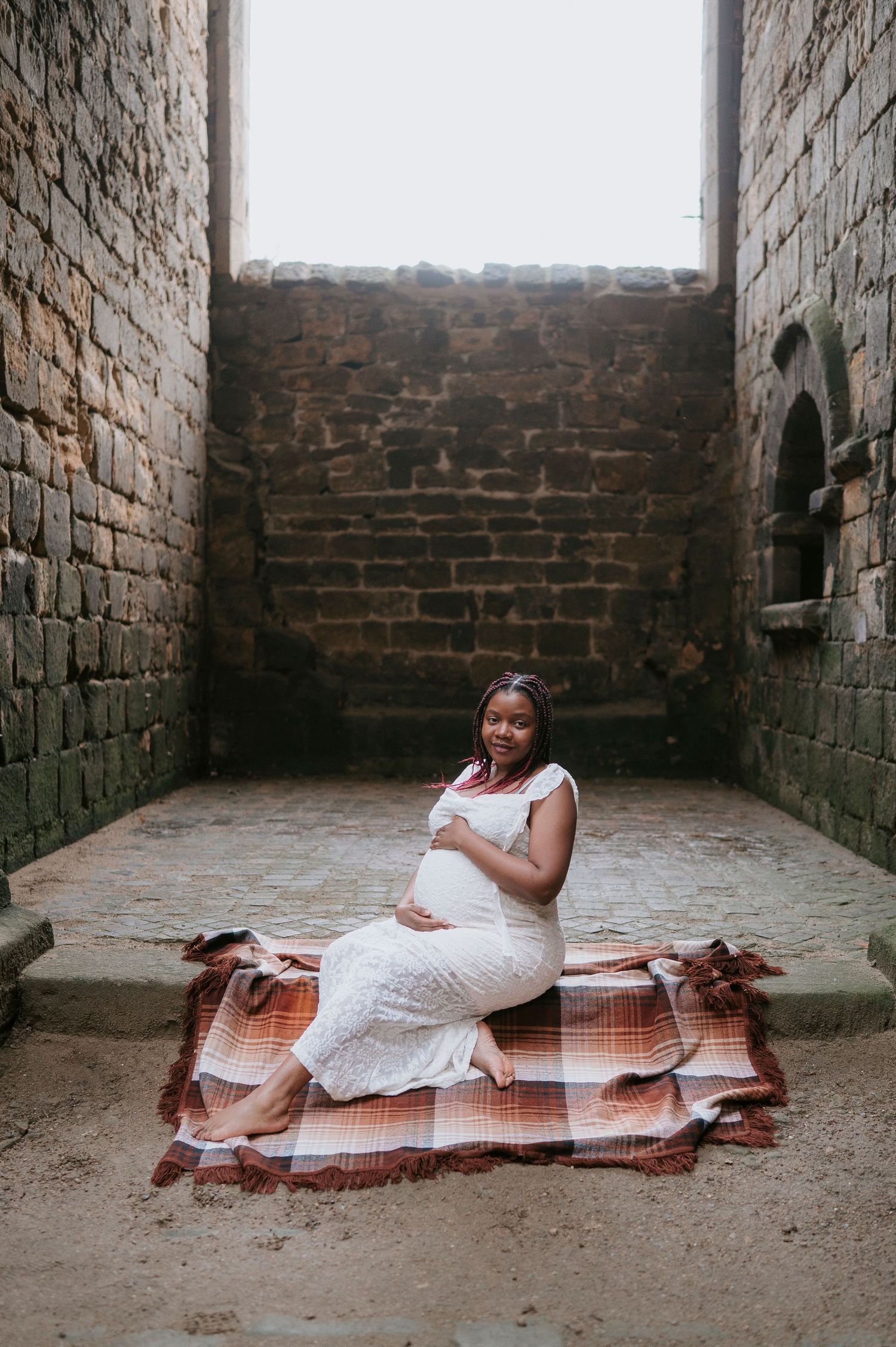 A series of maternity photos showing a woman in a white dress reclining on a brown blanket against rustic brick walls.