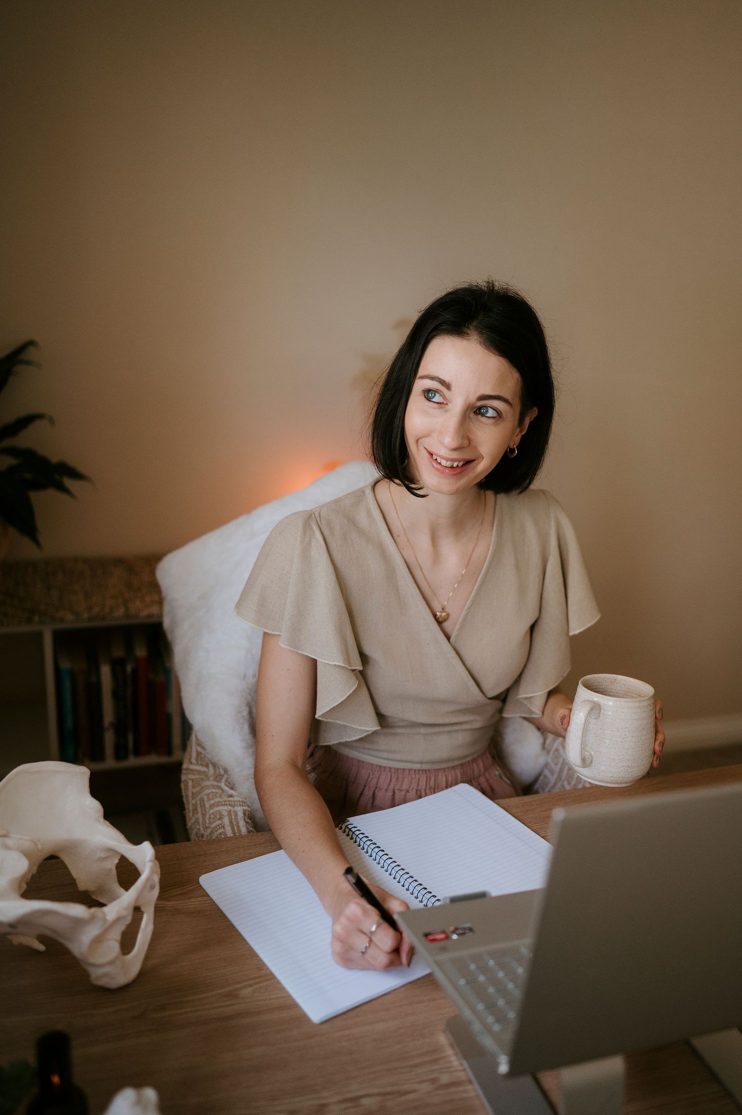 A person sits at a wooden desk writing in a notebook while holding a white coffee mug in warm lighting.
